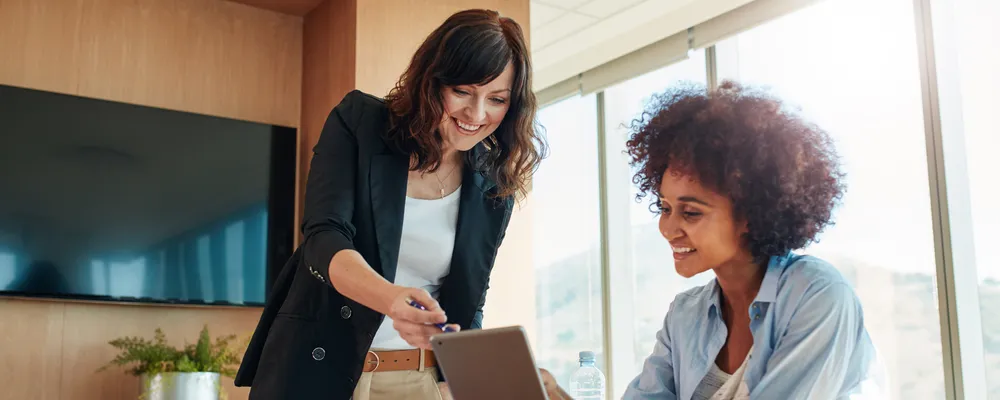 One woman holding a tablet, with another helping, smiling, showing how to use it.